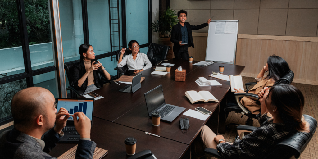 A group of people are sat in a boardroom conversing and working. One man is stood at the head of the table in front of a whiteboard presenting.