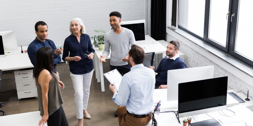 A group of people are stood in an office space conversing and working.