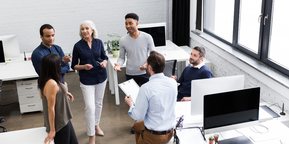 A group of people are stood in an office space conversing and working.