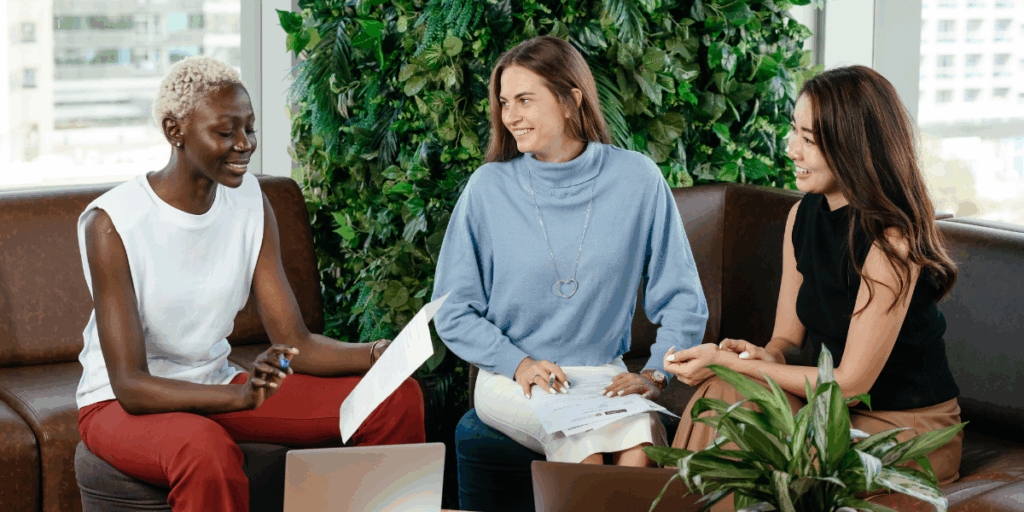 Three women are sat next to one another in a well-lit room conversing and reviewing papers.