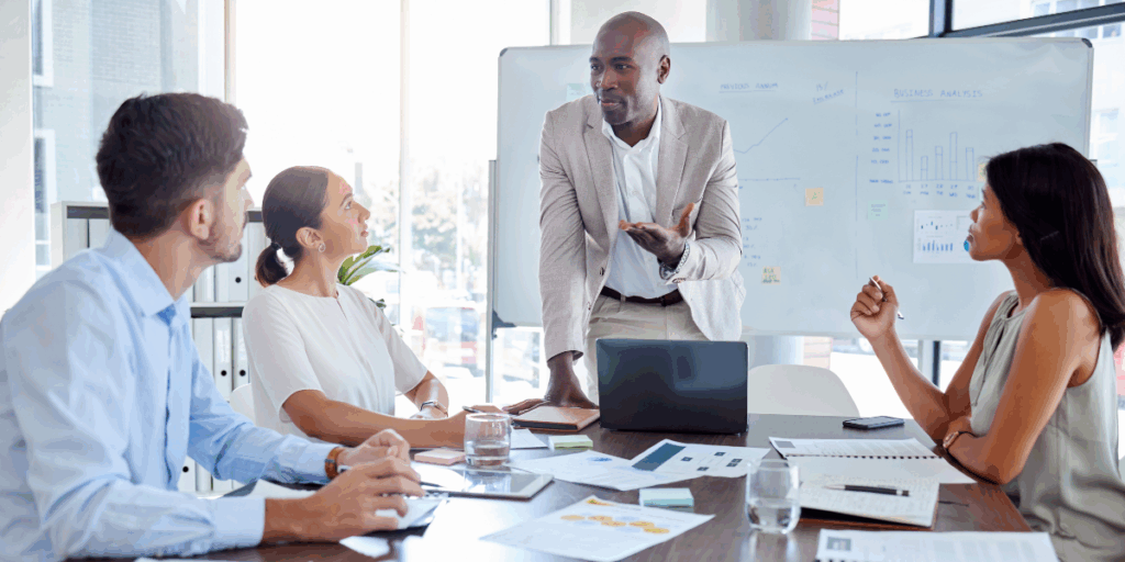 A man is stood at the front of a boardroom table presenting to two people.