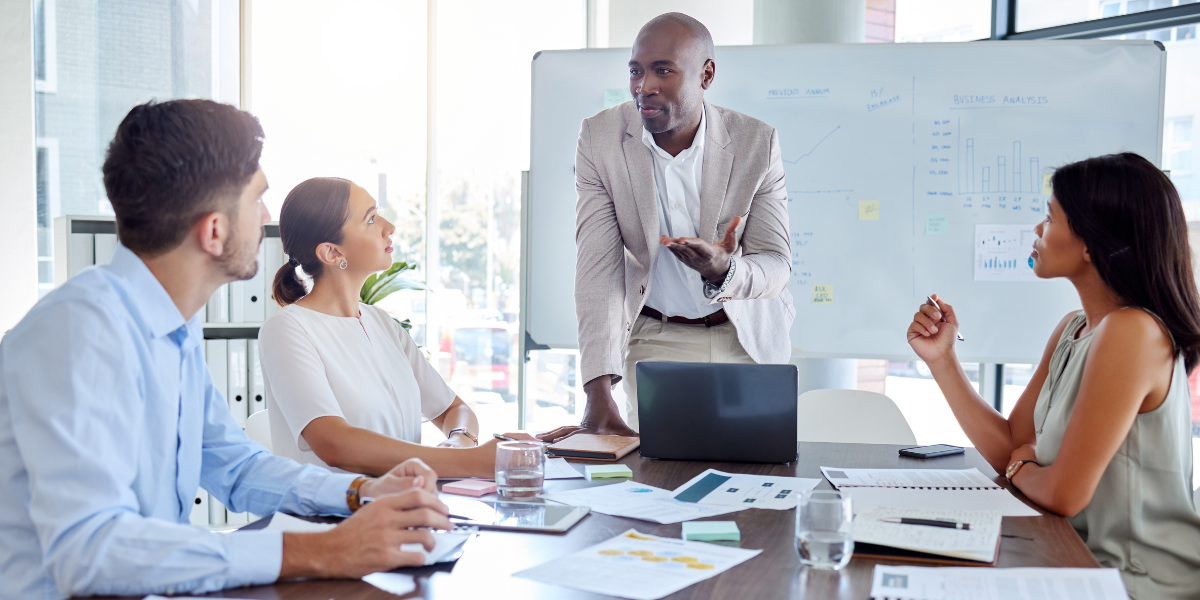 A man is stood at the front of a boardroom table presenting to two people.