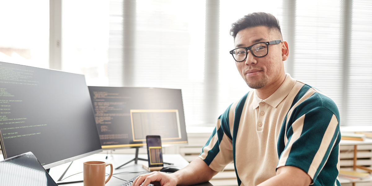 A man is sat at a desk in front of two monitors displaying HTML code.