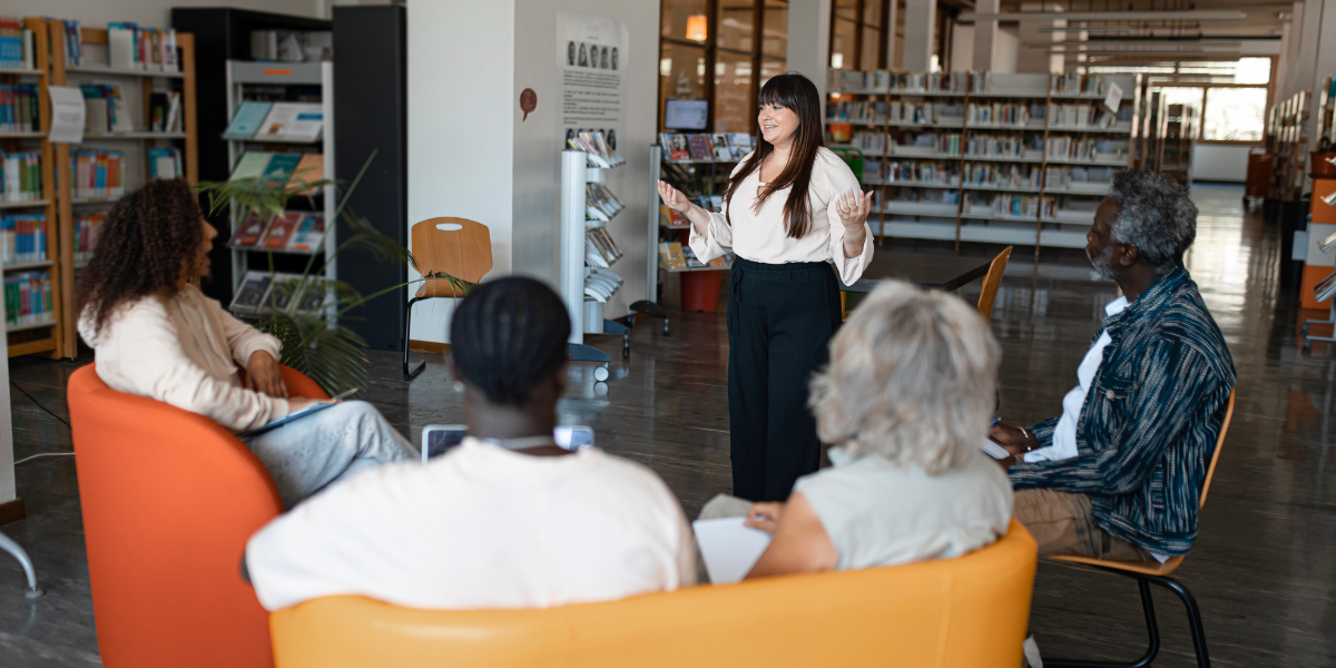 A woman is stood in front of a group of people presenting in a library.