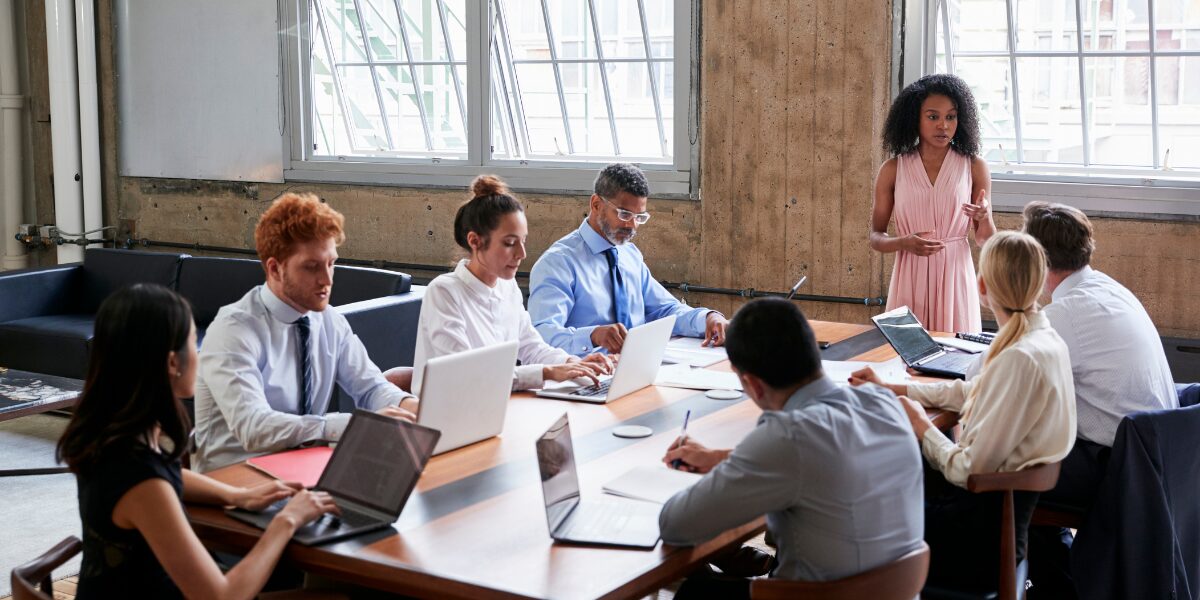 A woman is stood at the front of a boardroom table presenting to a group of colleagues.