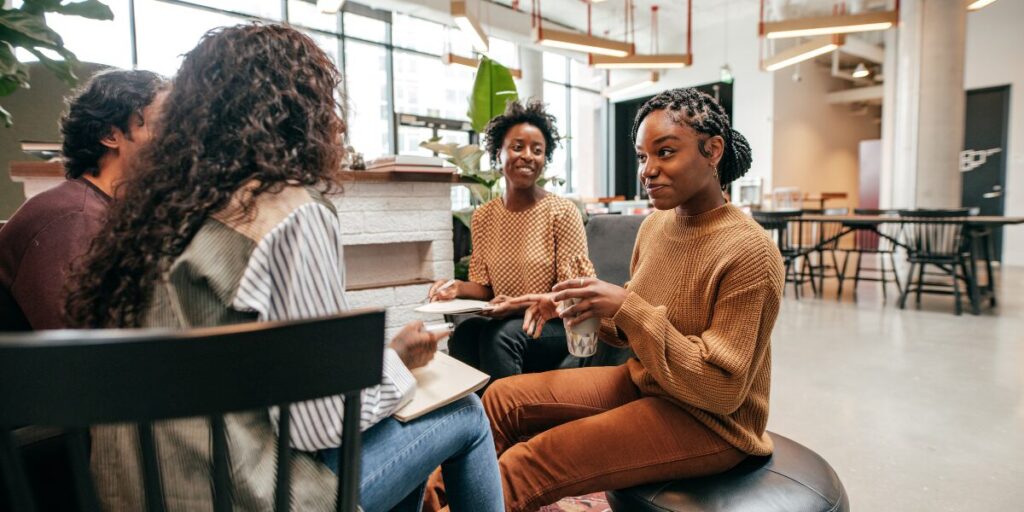 A group of people are sat in a well-lit office space conversing and laughing.