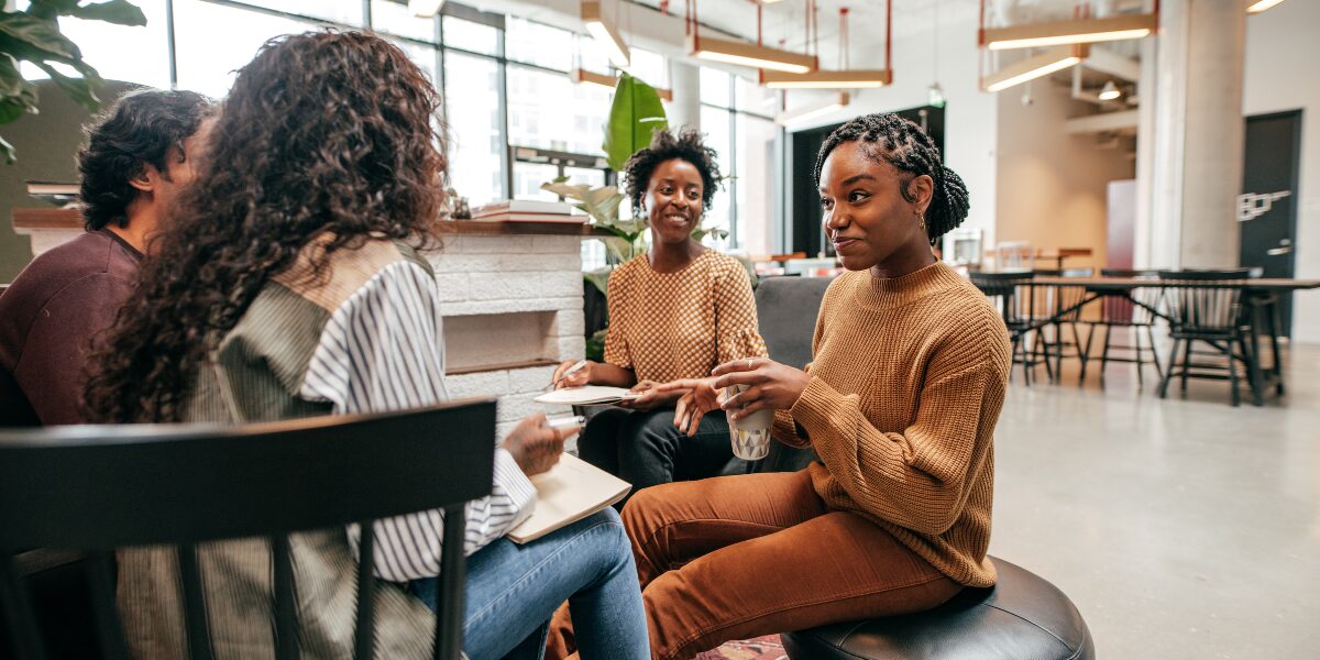 A group of people are sat in a well-lit office space conversing and laughing.