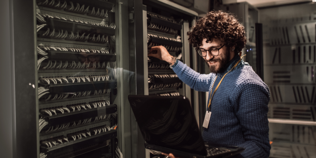A man is stood in an IT networking room working and holding a laptop.