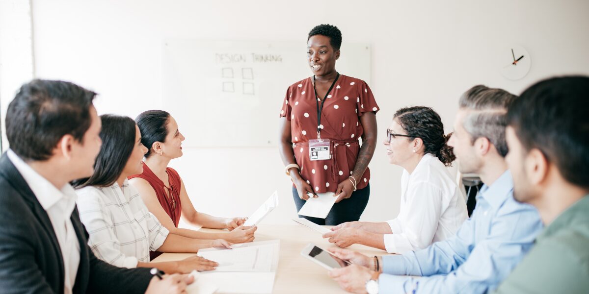 A woman is stood at the front of a boardroom holding a piece of paper and presenting to a boardroom of people.