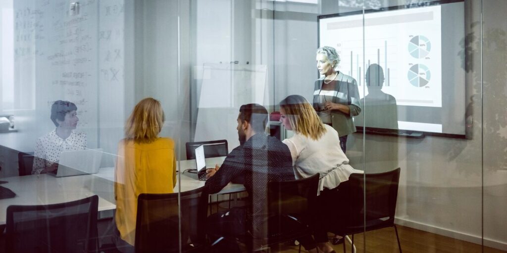 A woman is stood at the front of a boardroom presenting to a group of people.
