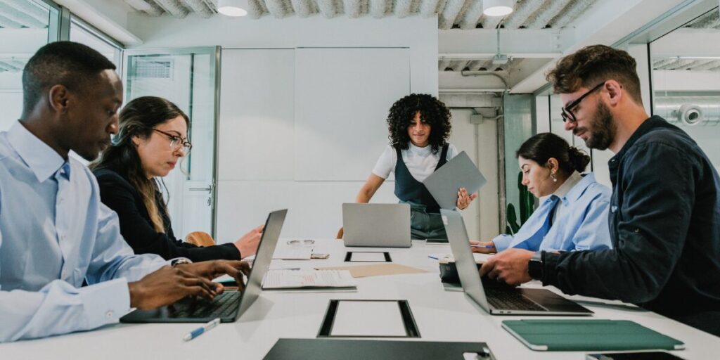 A woman is stood at the head of a boardroom table. A group of people are sat at the table.