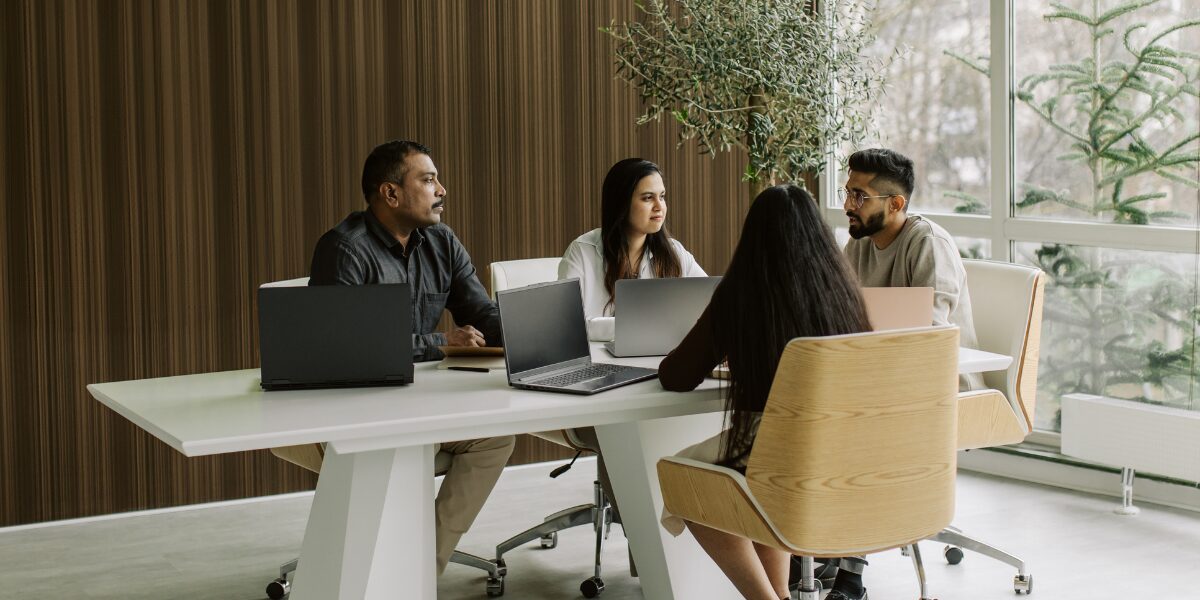 A group of people are sat at a boardroom table working and conversing.