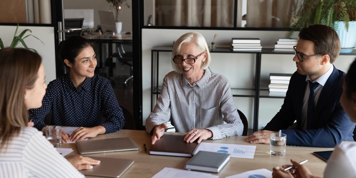A group of people are sat at a boardroom table conversing and working on a project.