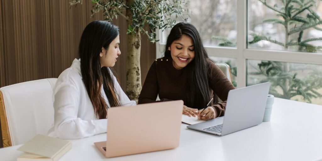 Two women are sat next to one another working on a document in a well-lit office space.