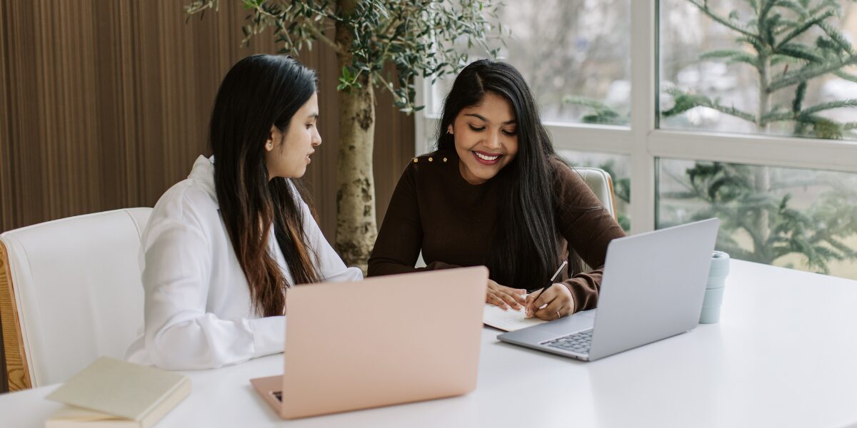 Two women are sat next to one another working on a document in a well-lit office space.
