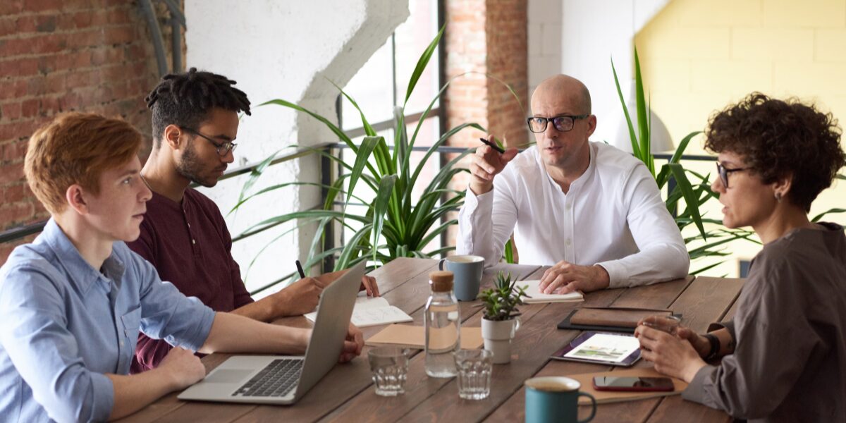 A man is sat at the head of a boardroom table speaking to his team.