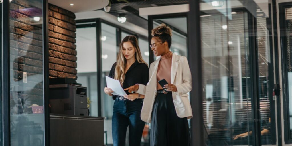 Two women are walking side-by-side in an office space reviewing a document.