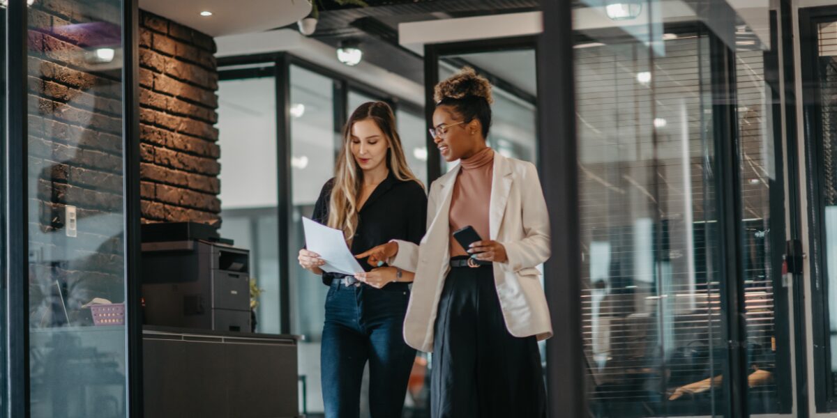 Two women are walking side-by-side in an office space reviewing a document.