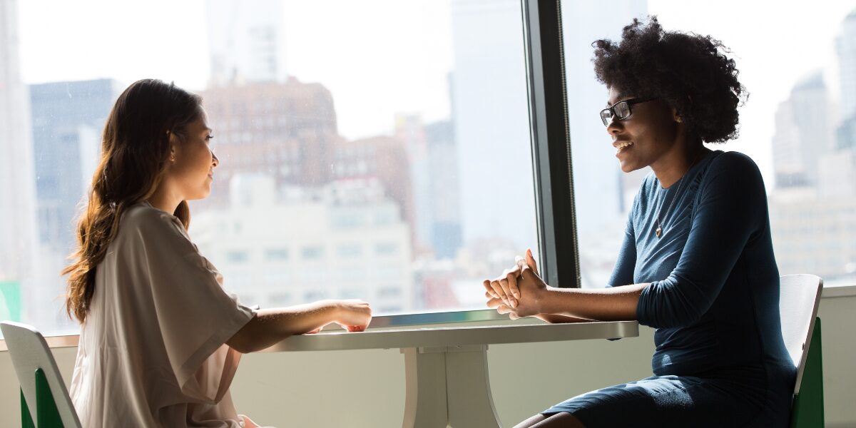 Two women are sat at a high-top table in front of a set of windows conversing.