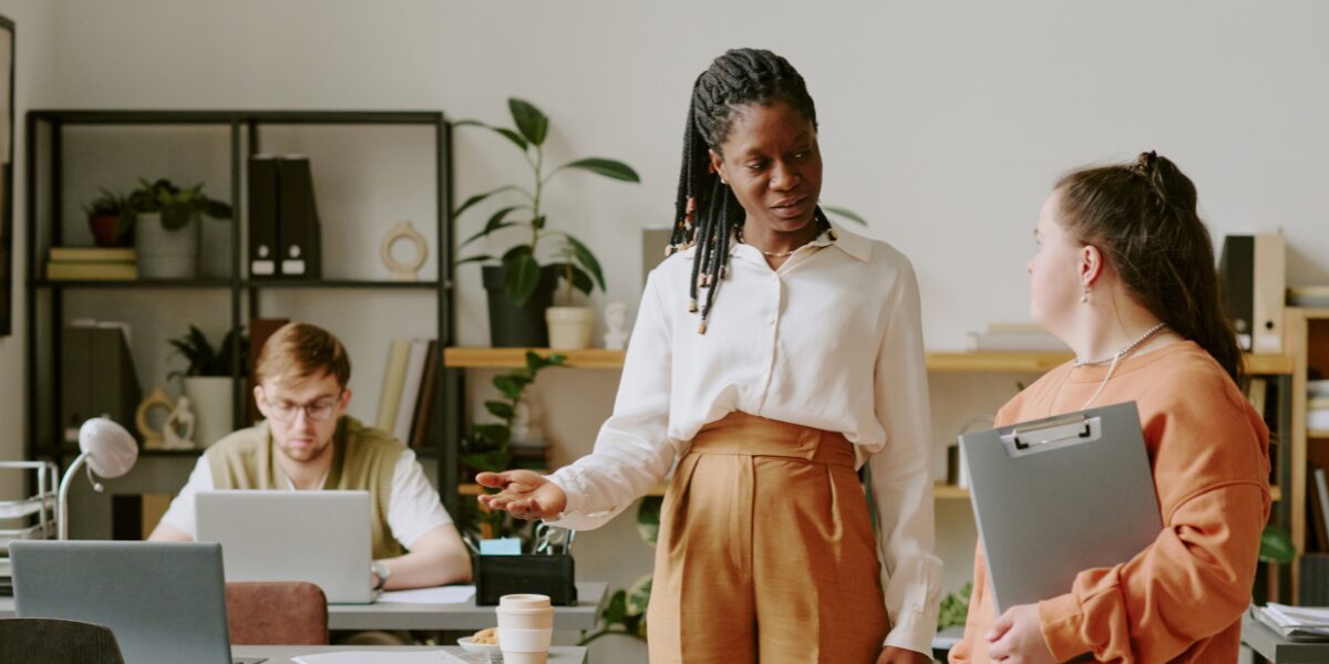 Two women are stood in an office space conversing and one is gesturing to enter a different room.