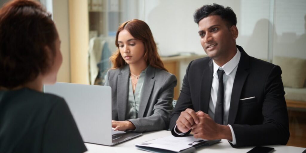 Three people are sitting at a boardroom table. The man is interviewing a job candidate.