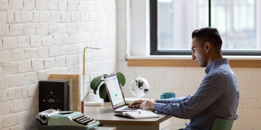 A man is sat at his desk working on his laptop in a well-lit office space.