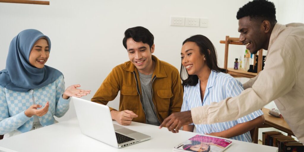 Four people are working in an office space. They are gathered around a laptop in a well-lit room conversing and reviewing something on the laptop.