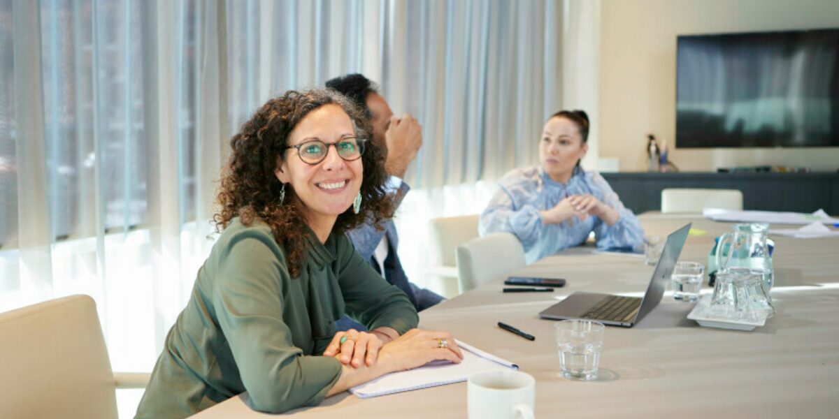 A woman is sat at a boardroom table smiling at the camera. While two co-workers are sat working in the background.