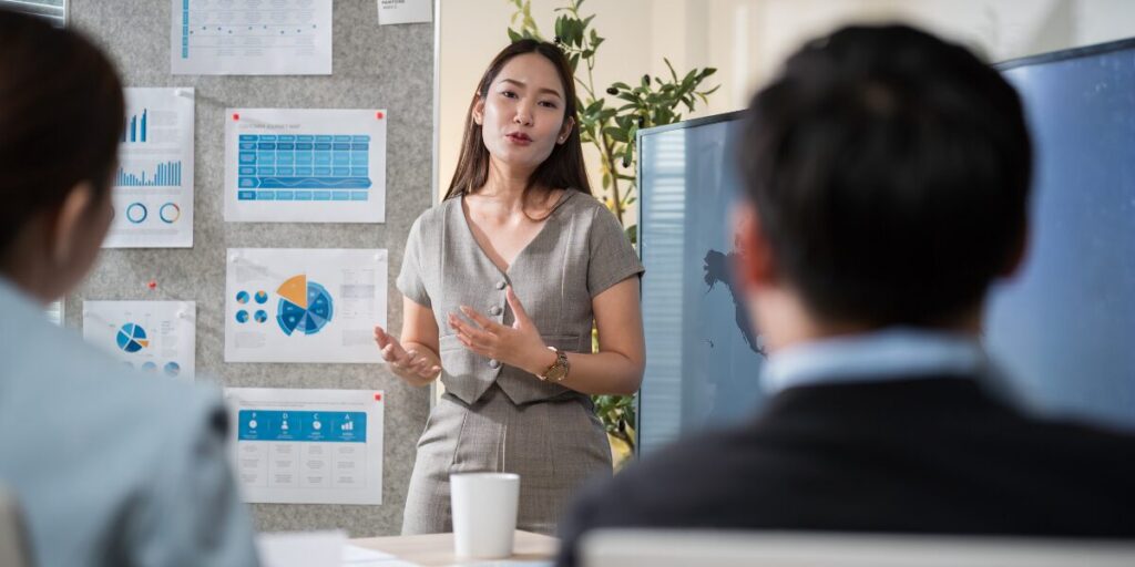 A woman is stood in front of a bulletin board presenting to her colleagues in a boardroom.
