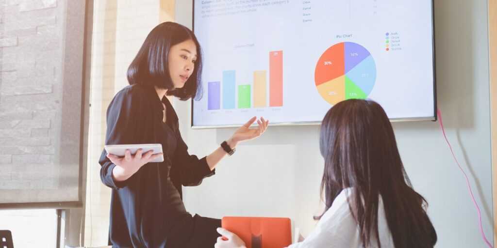 A woman is sat on a desk holding a tablet and pointing at a slide deck projected on the wall. The woman is presenting to her colleagues.