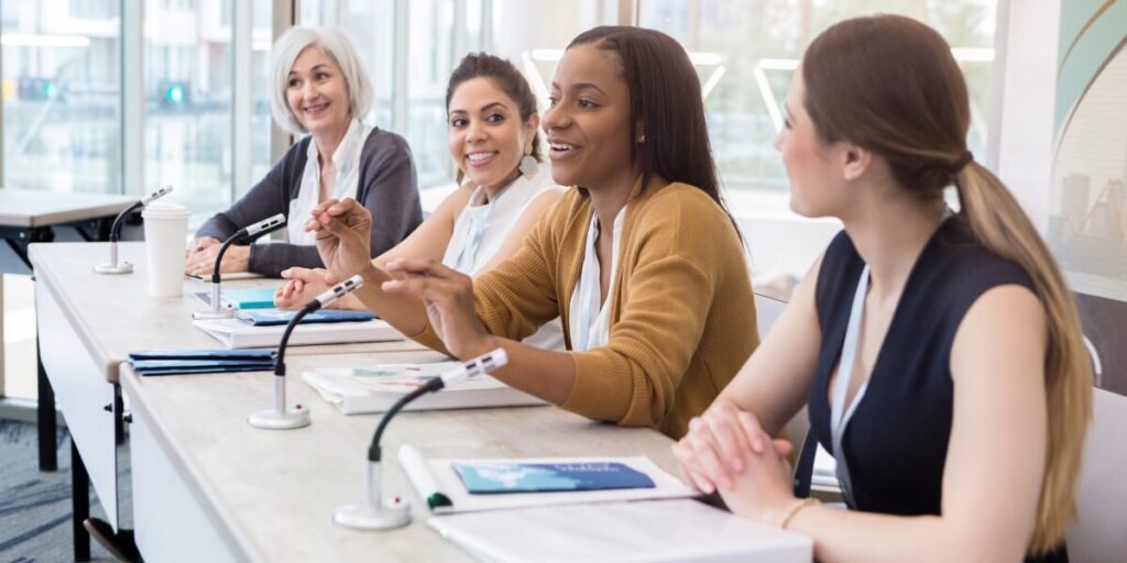 Four women are sat at the head of board room table speaking to an audience.