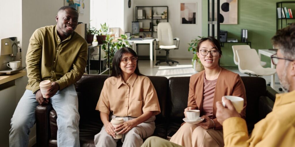 Four people are sat in an office space conversing. Three people are sat on a couch and one person is sat on a chair.