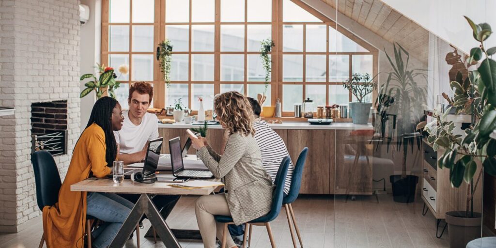 Four people are sat in a boardroom working and conversing at a shared table.