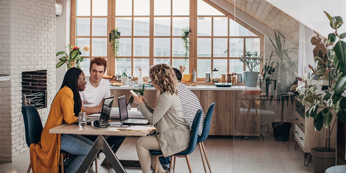 Four people are sat in a boardroom working and conversing at a shared table.