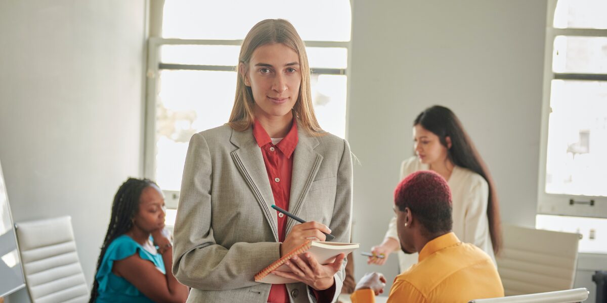 A woman is stood facing the camera in an office space, and holding a note pad. There are three co-workers sat behind them.