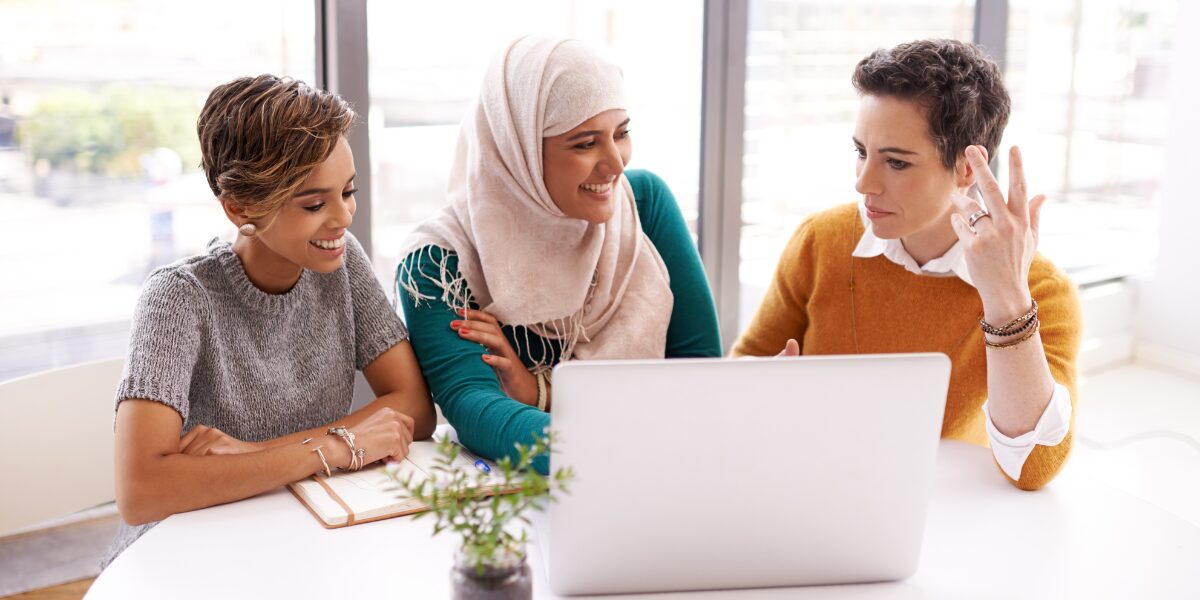 Three people are sat at a boardroom table collaborating and conversing while reviewing a laptop screen.