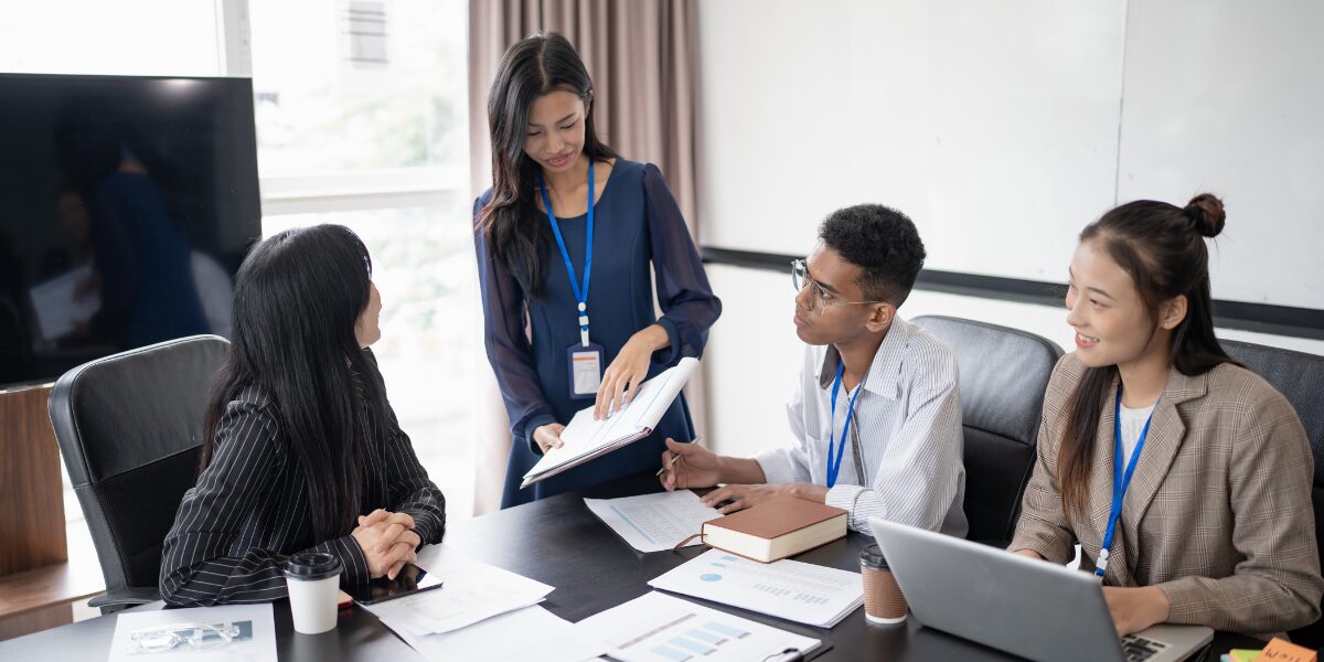 A woman is stood presenting and reviewing a document. Three people are sat at a boardroom table listening.