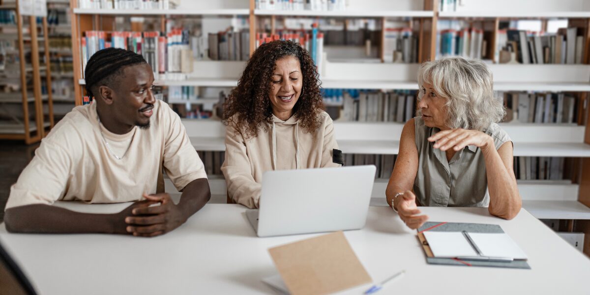 Three people are sat in an office space collaborating, and reviewing a laptop.