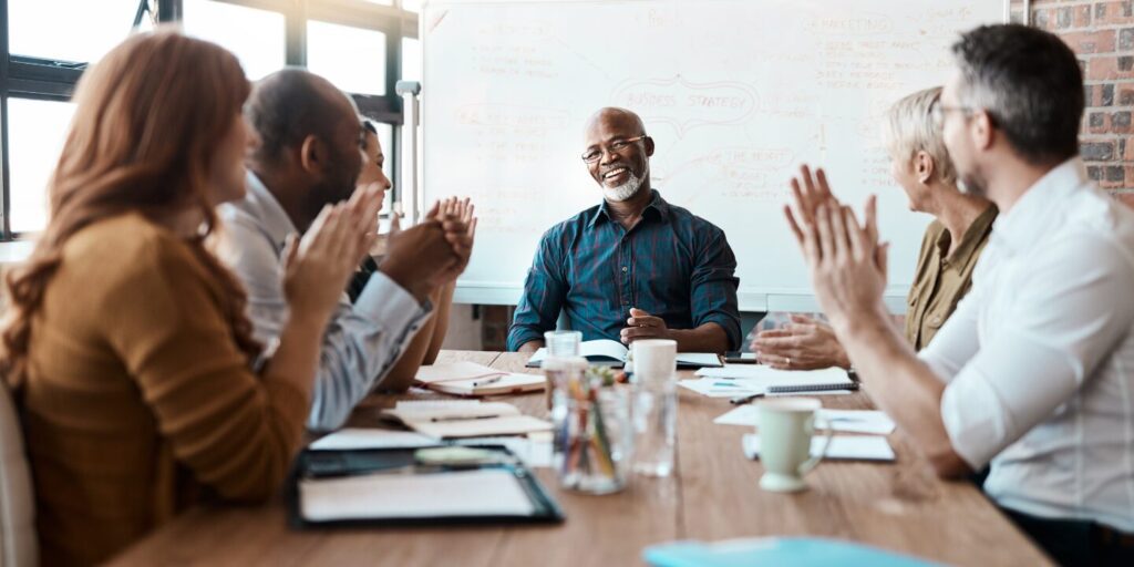 A man is sat at the head of the boardroom table smiling, and colleagues are lining the boardroom table clapping in celebration.