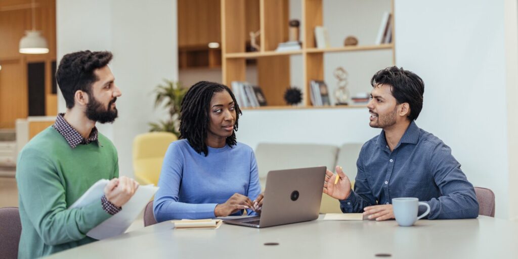Three people are sat at a boardroom table in a well-lit room conversing. One person has their laptop open on the table.