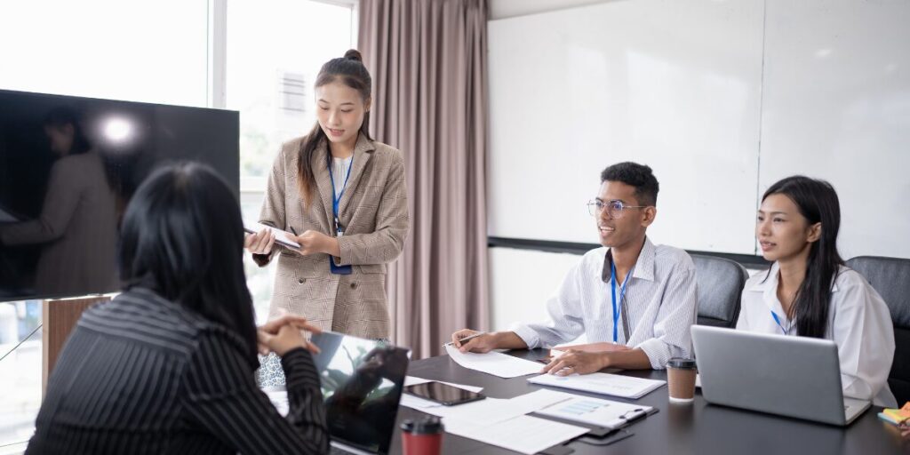 A woman is stood presenting and reviewing a document. Three people are sat at a boardroom table listening.