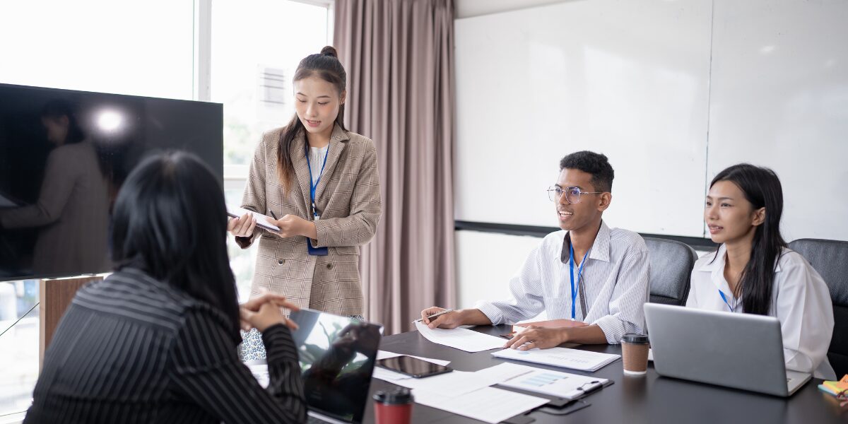 A woman is stood presenting and reviewing a document. Three people are sat at a boardroom table listening.