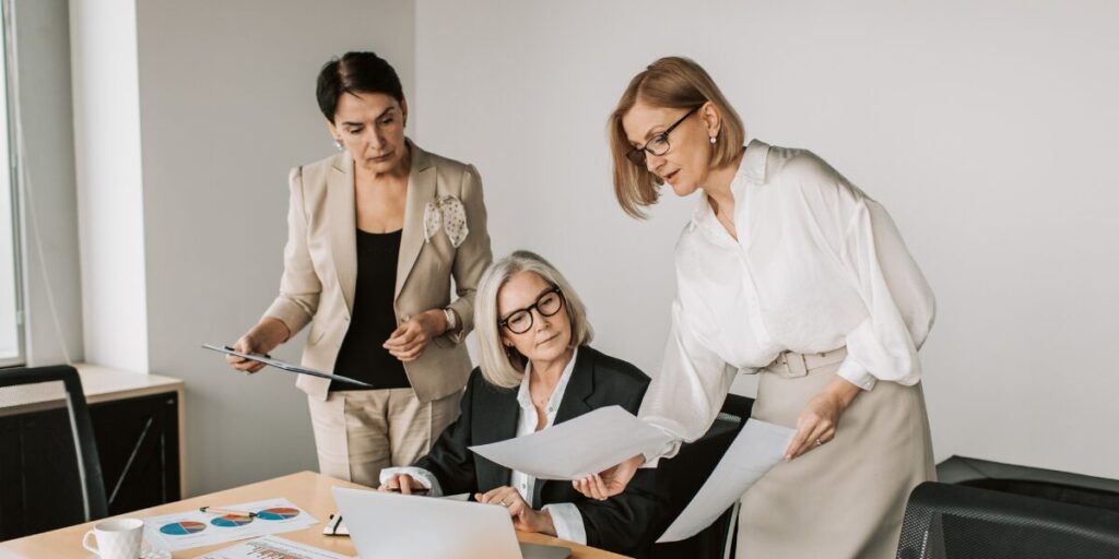 Three women are working in a boardroom. One woman is sitting reviewing documents and the other two are standing.