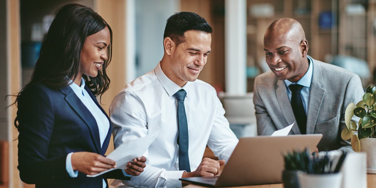 Three people are gathered around a boardroom table reviewing documents and conversing.