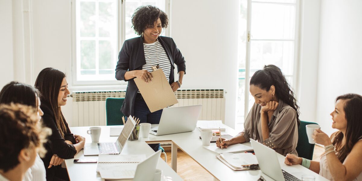 A woman is stood at the front of a boardroom table presenting to a team.