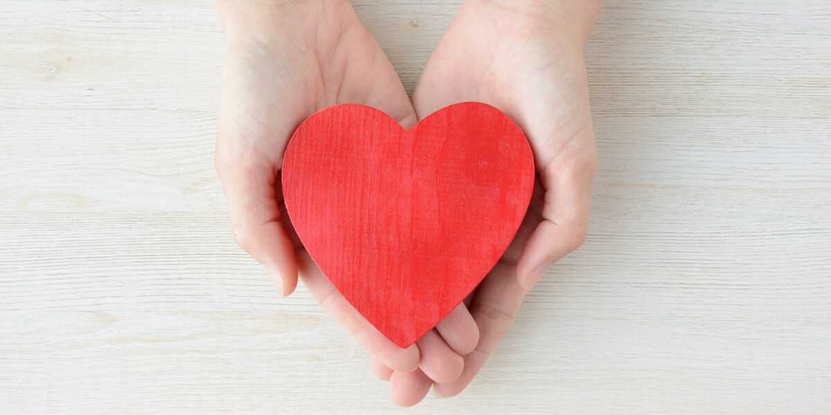 Two hands holding a red heart on a white table.