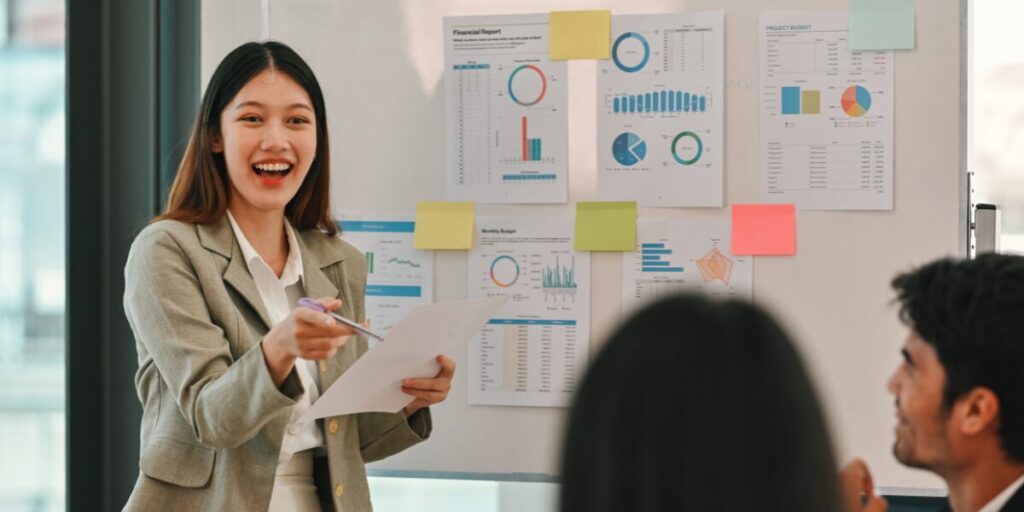 A woman is stood at the front of a boardroom presenting to colleagues and reviewing stats.