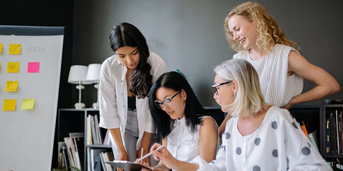 Four women are working in an office space conversing and collaborating.