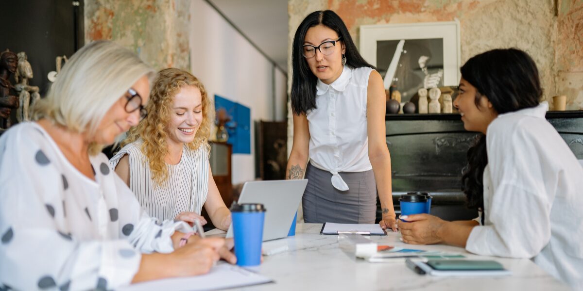 A woman is stood at the head of a boardroom table presenting to three women seated at the table.