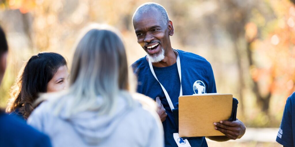 A man in a volunteer uniform wearing a lanyard is shaking hands with new volunteers.
